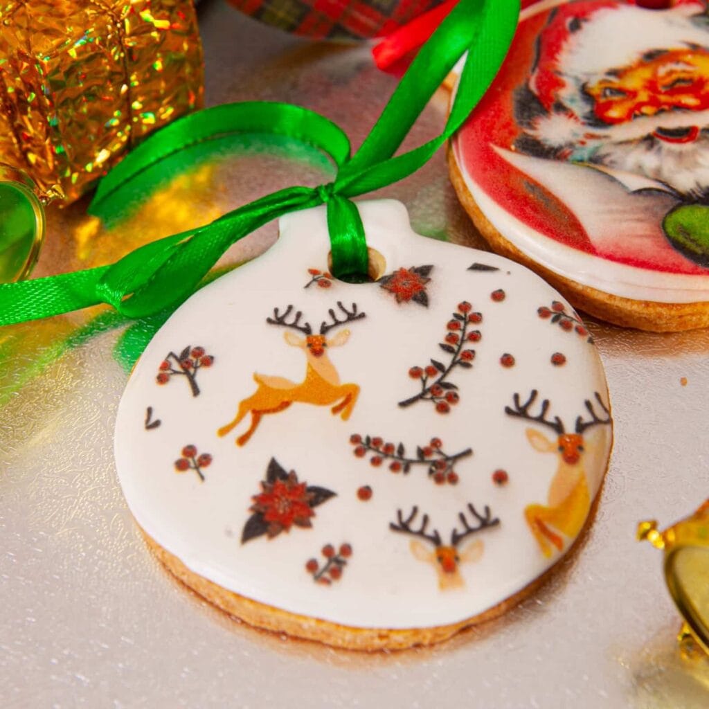 Galleta de Navidad en forma de bola con renos y flores de pascua impresos, atada con un lazo verde