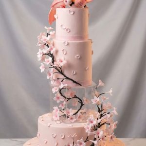 Tarta de boda rosa de tres pisos decorada con ramas de flores de cerezo y figura de pajaritos enamorados con velo en la cima.